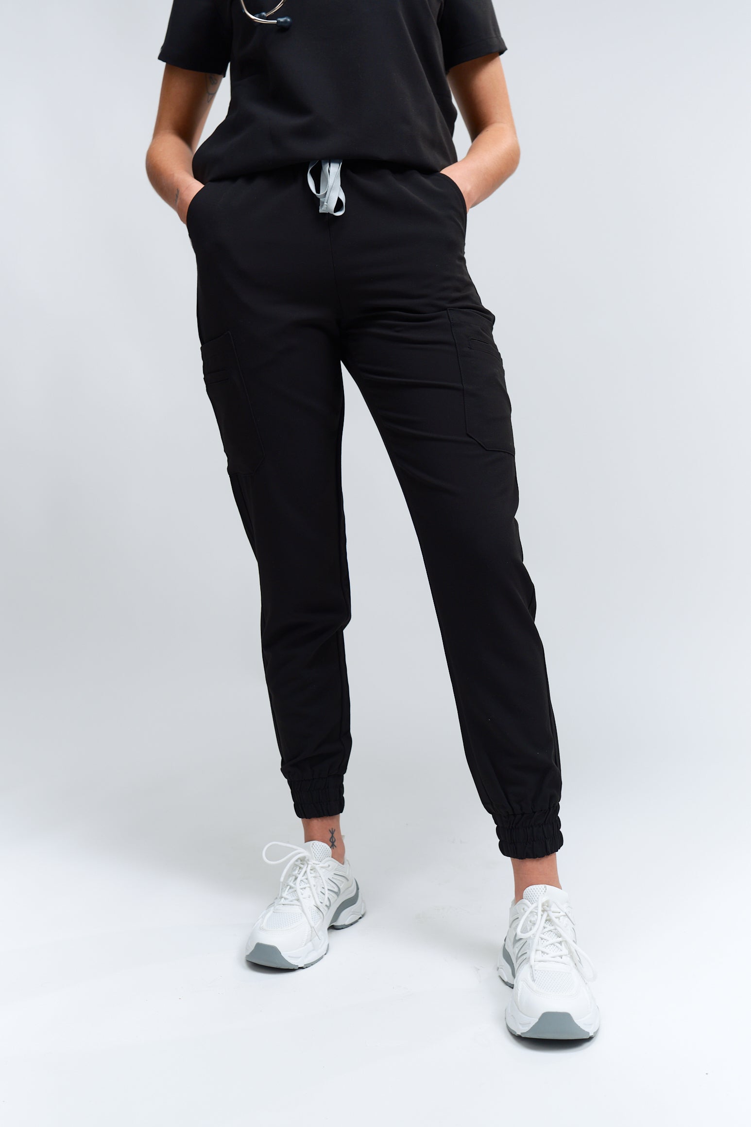 A person wearing black jogger scrub pants with multiple pockets and a drawstring waist, paired with a black scrub top and white sneakers.